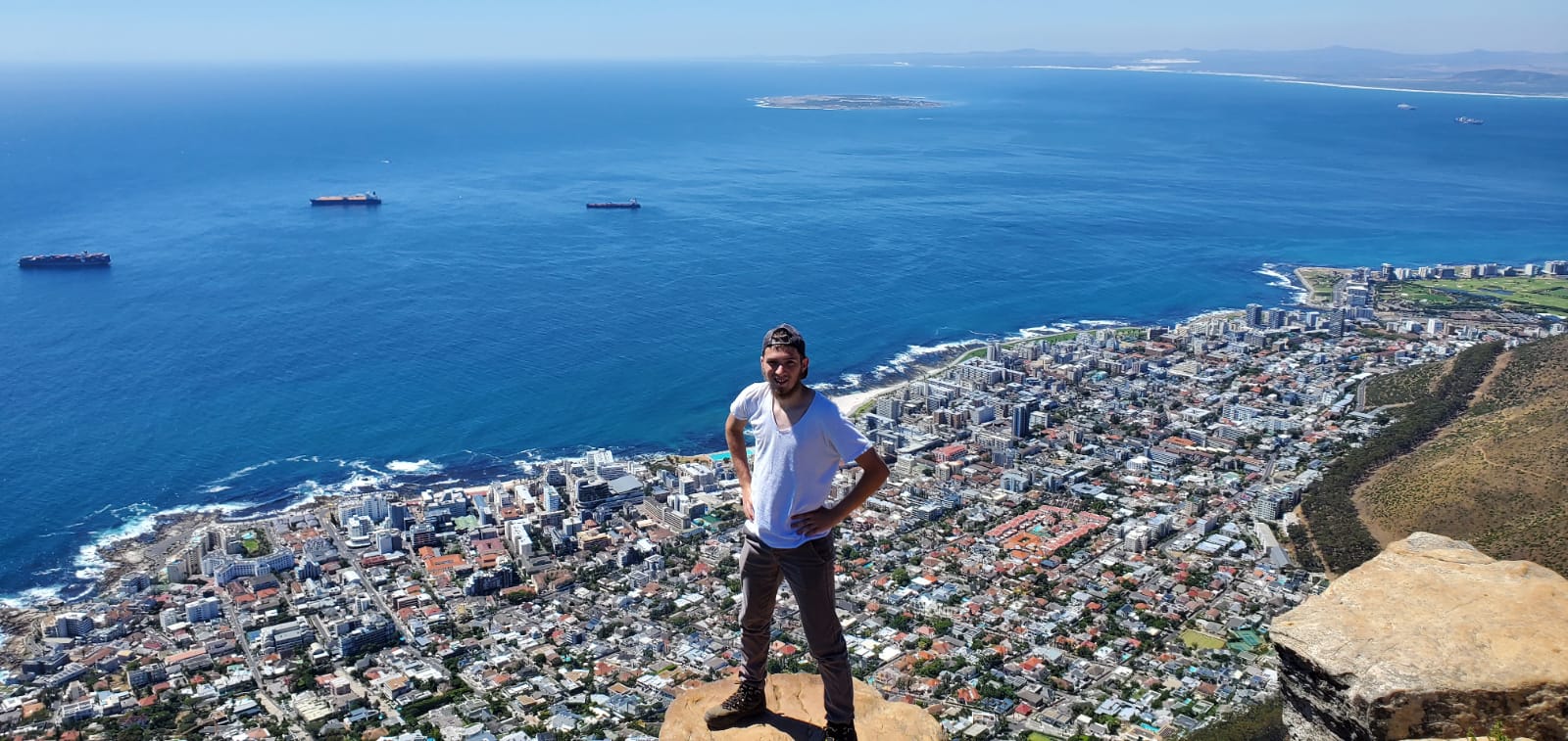 A man in a white t-shirt and cap stands on a cliffs edge overlooking Cape Town.
