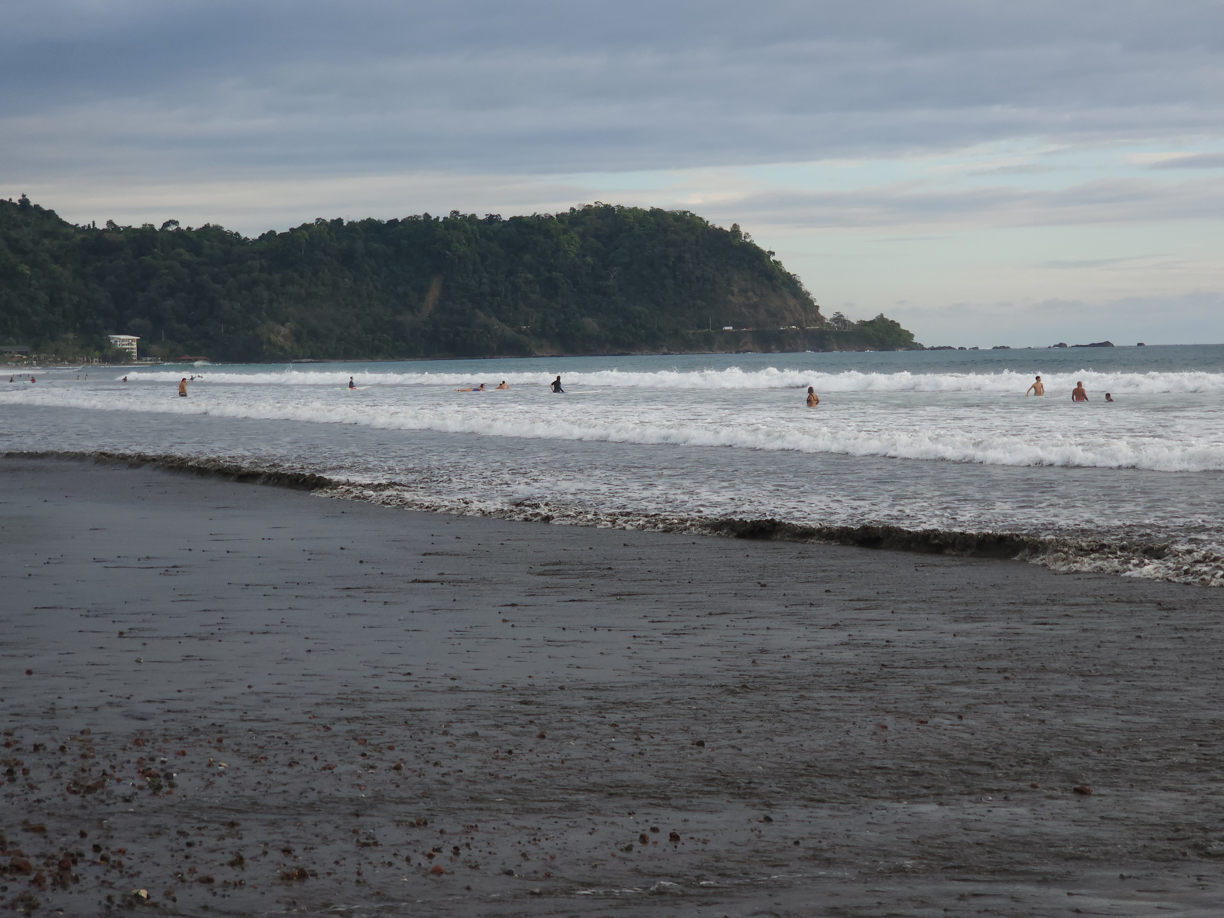 People frolic in the short waves at a black sand beach on a cloudy day