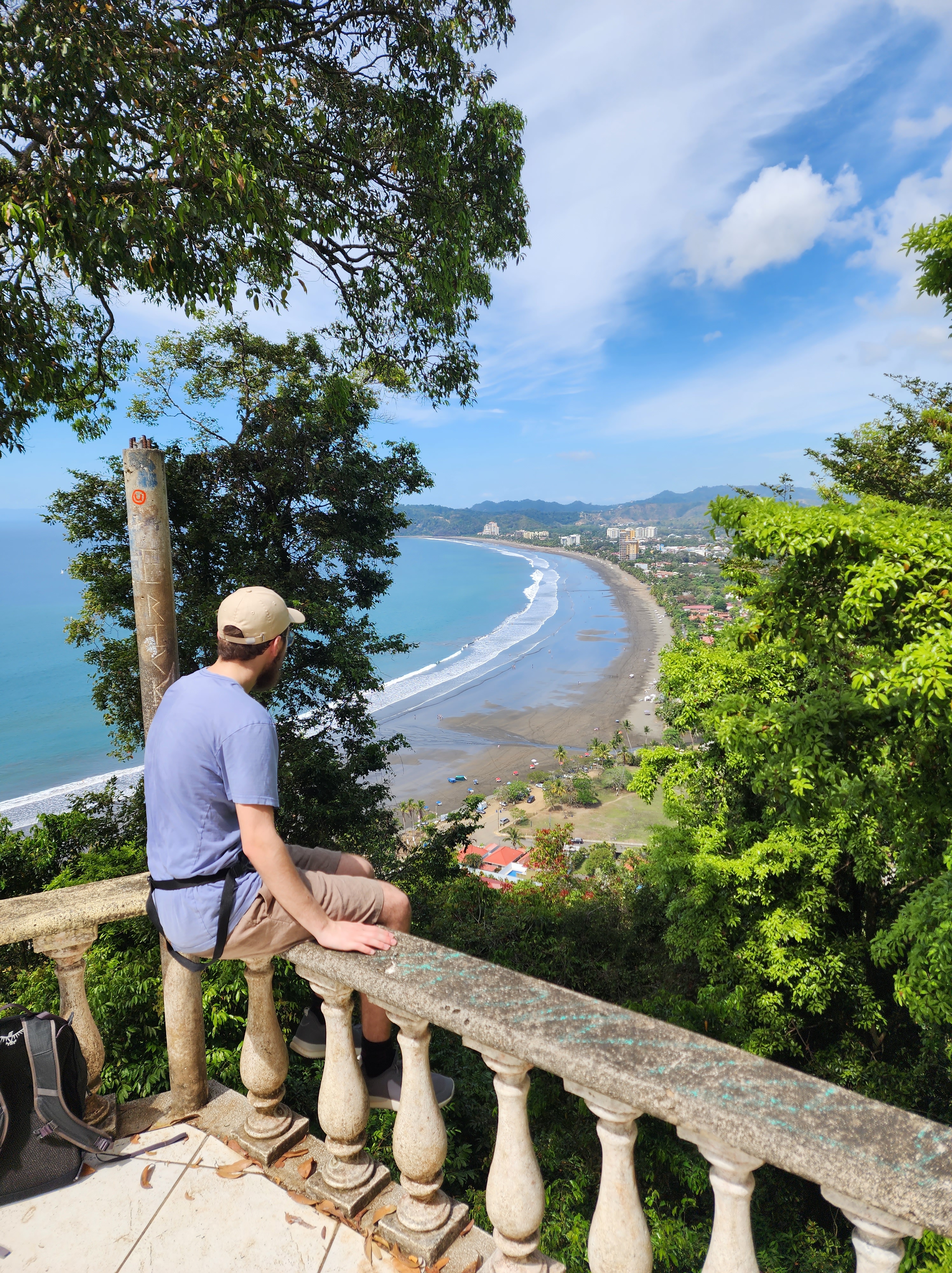 A man in a blue shirt and beige cap sits on a stone railing overlooking an expansive view a long beach out ahead