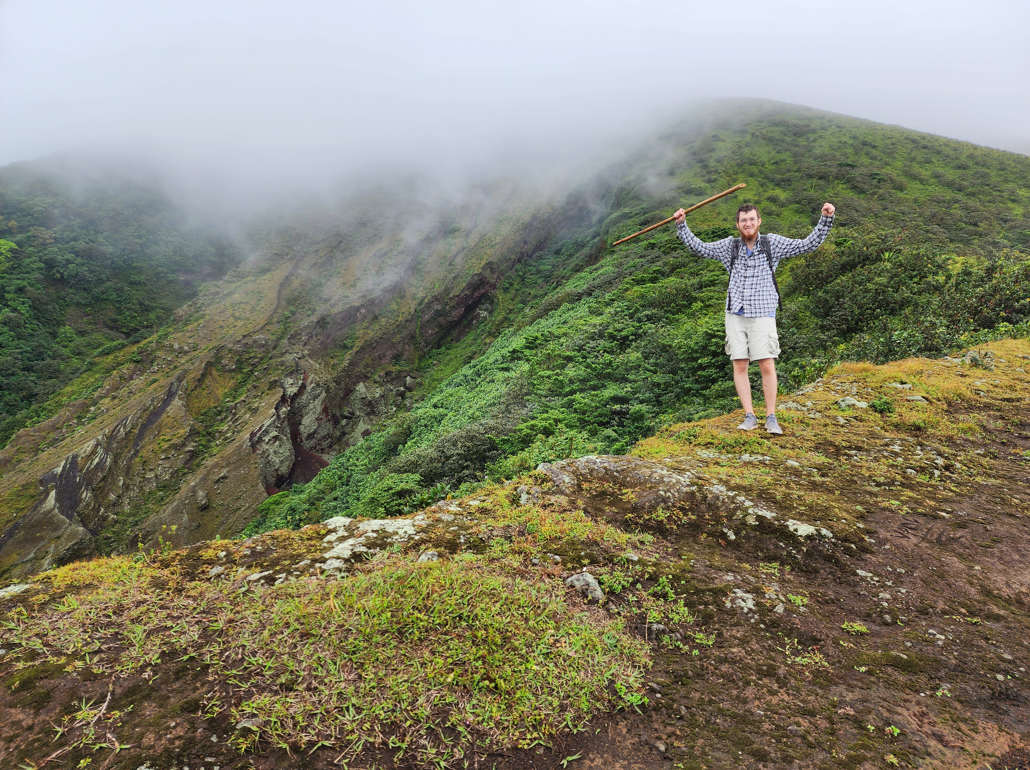 A man lifts his hiking staff in triumph on a mossy green mountainside