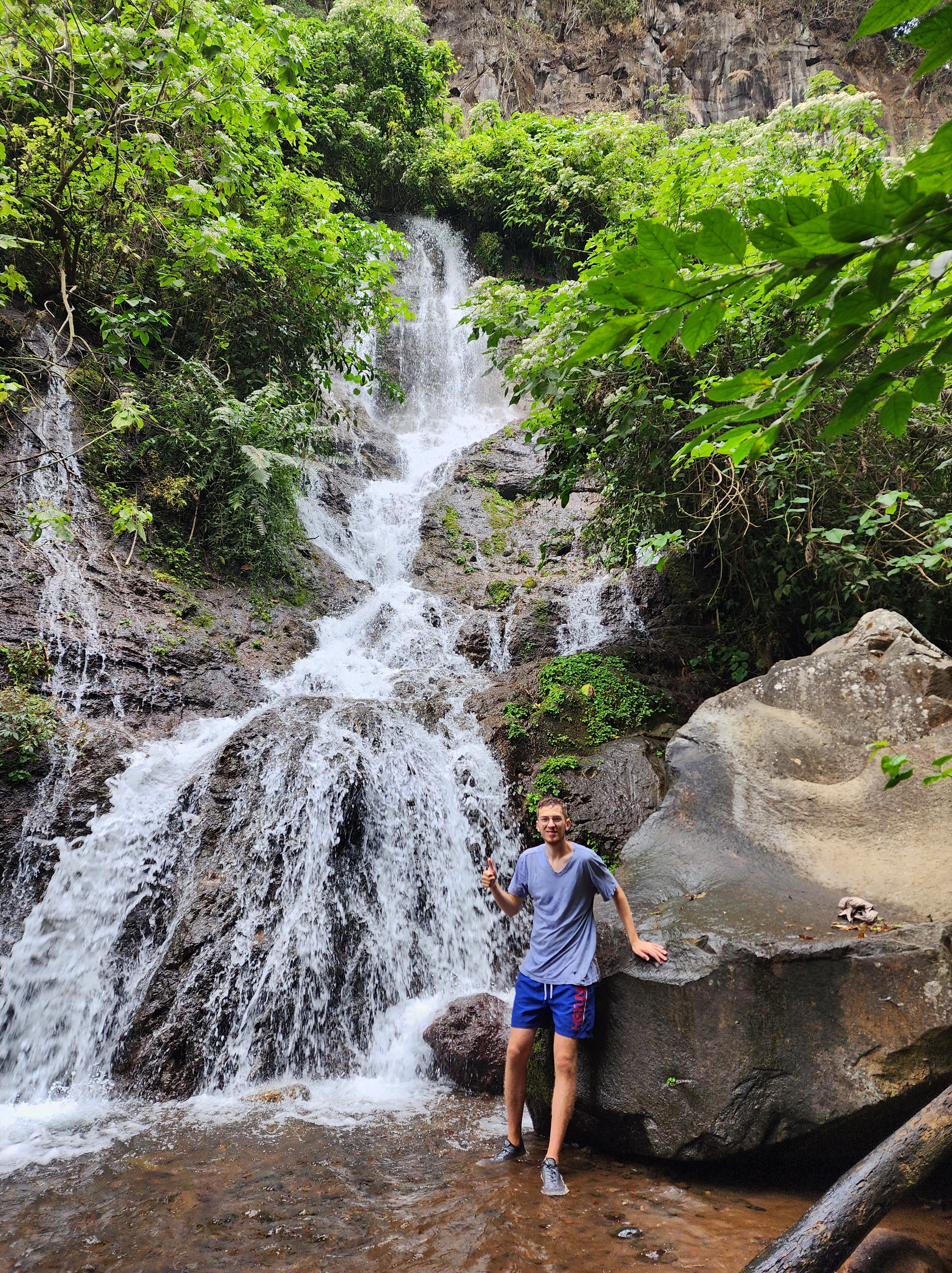 A man in a blue t-shirt and swim trunks gets drenched while standing next to a tiered waterfall in the jungle