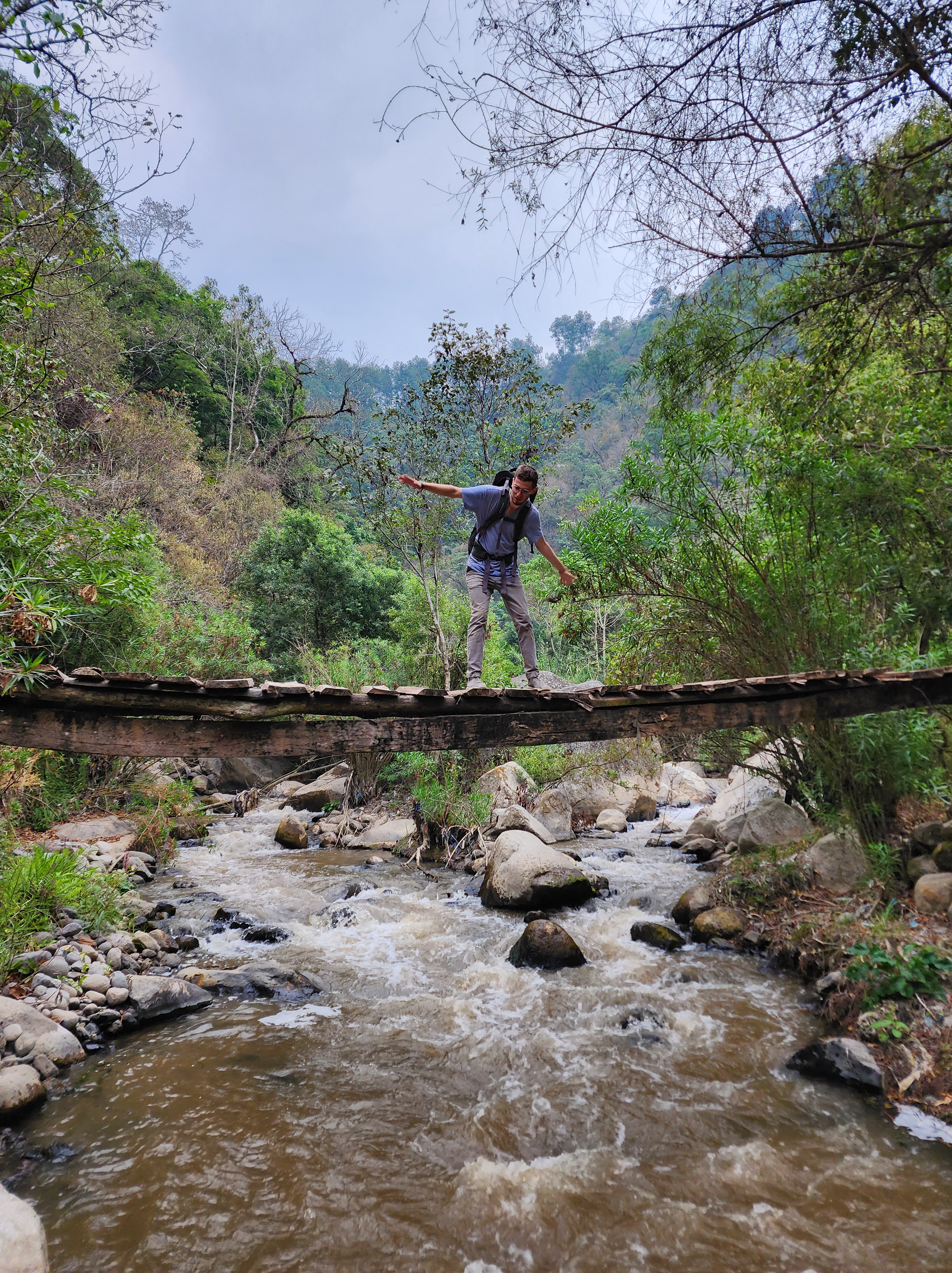 A man precariously balances on a wooden bridge without safety rails that crosses a rocky river