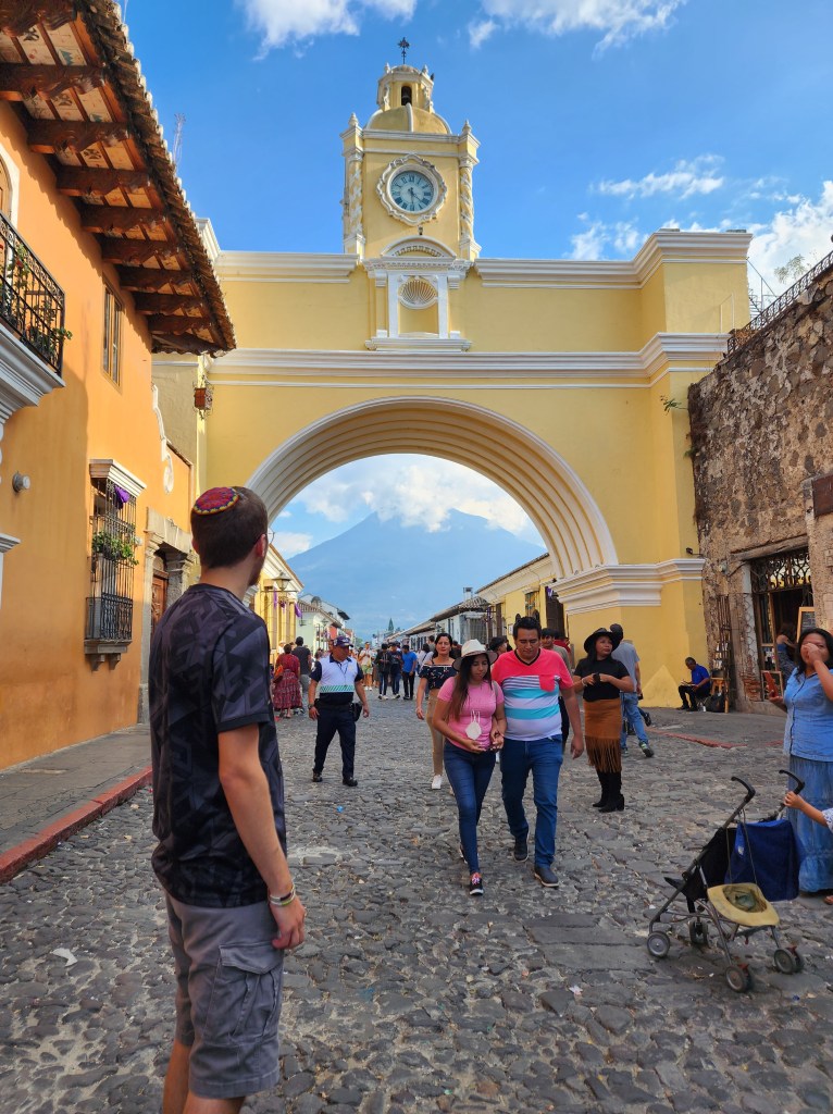A man stands on a cobblestone street, looking at a yellow and white arch with a clock in the centre. In the view beyond the arch is a large mountain right outside of the city.