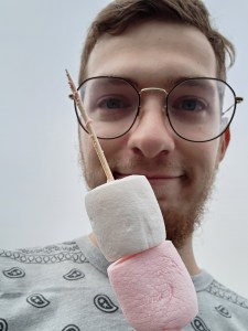 A man makes a weird face while holding up a skewer with two marshmallows on it; one white and one pink