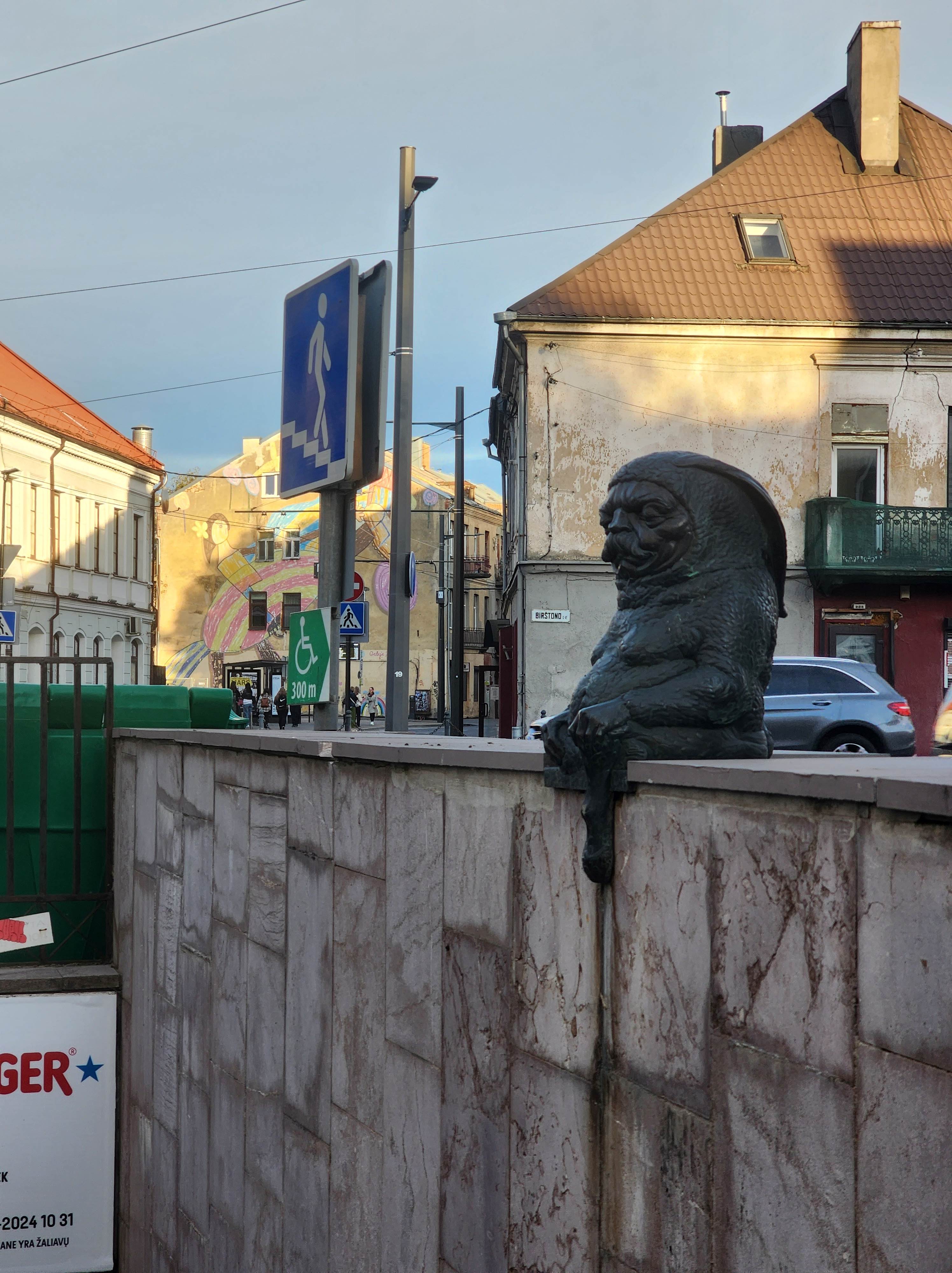 A small sculpture of a gnome like creature sitting on the edge of a subway station wall
