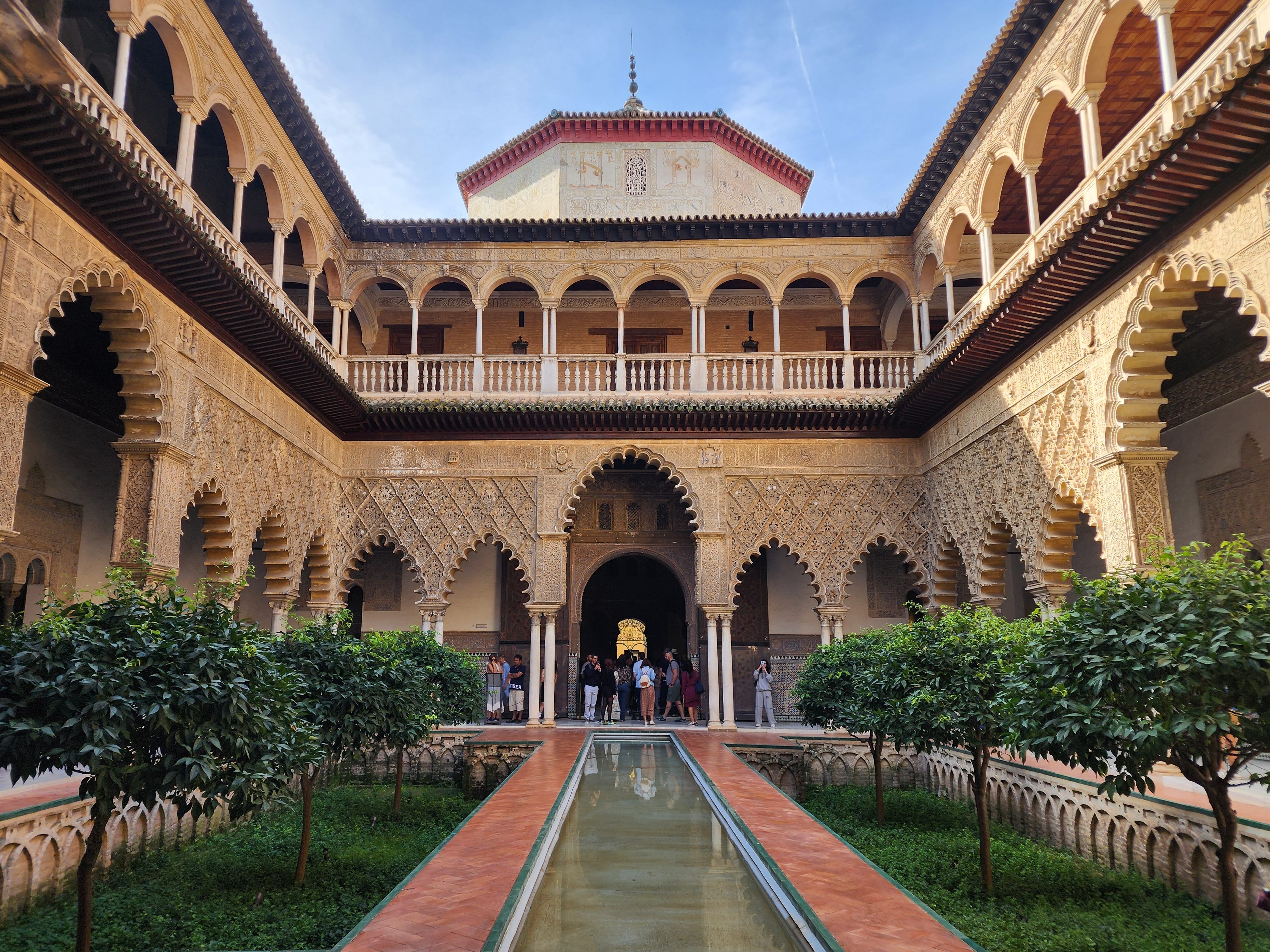 The main patio in the Royal Alcazar of Seville. The perfectly centred rectangular pool is lined by pretty trees and Mudejar architecture