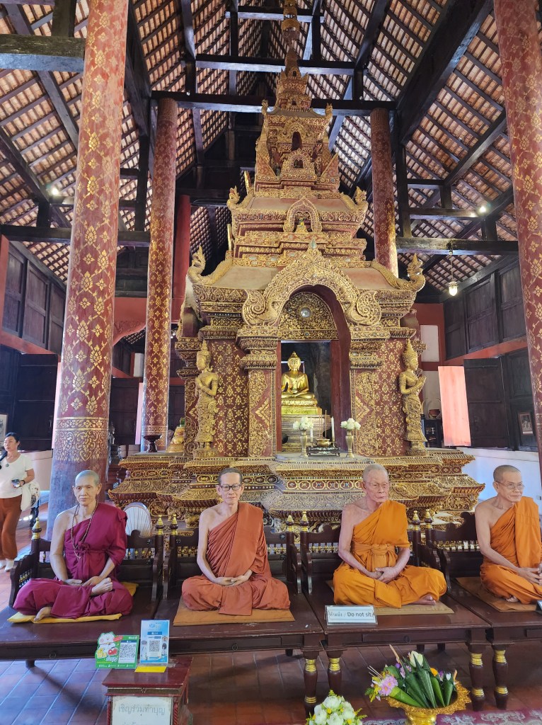 Five wax statues of meditating Thai monks dressed in orange, placed in front of a shrine.
