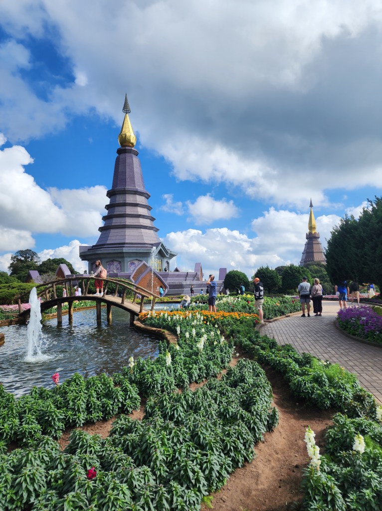 A pretty garden with a bridge over a clear pond, in front of two modern pagodas which are painted purple with gold spires.