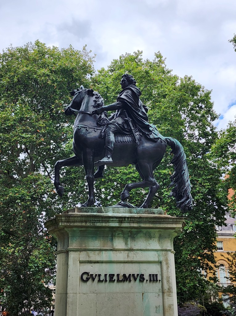 An iron statue of a king on his horse, on a pedestal in a leafy park.