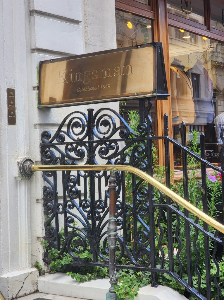 A gold sign saying "Kingsman; Established 1849" outside a tailors shop.