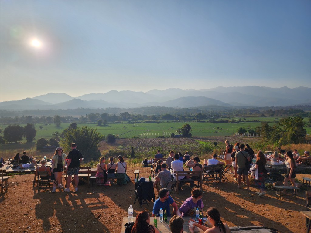 People sitting on benches and picnic blankets on a hill overlooking a vividly green field with mountains in the background. At Two Huts Cafe in Pai, Northern Thailand.
