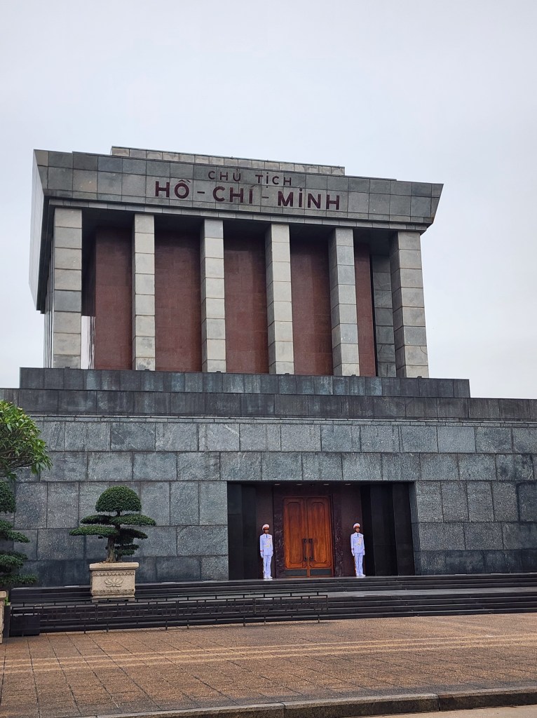 Two guards stand outside the entrance to Ho Chi Minhs Mausoleum. The building is large and with massive grey stones.